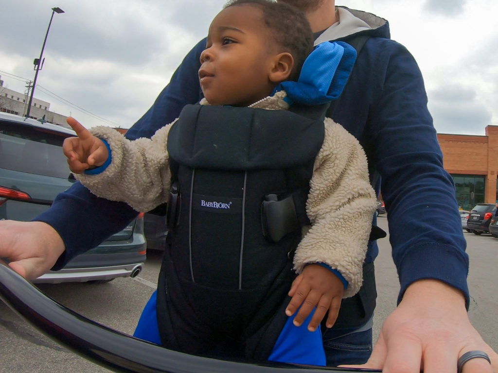 A kid in a baby Bjorn carrier carried by his dad who's pushing a shopping cart