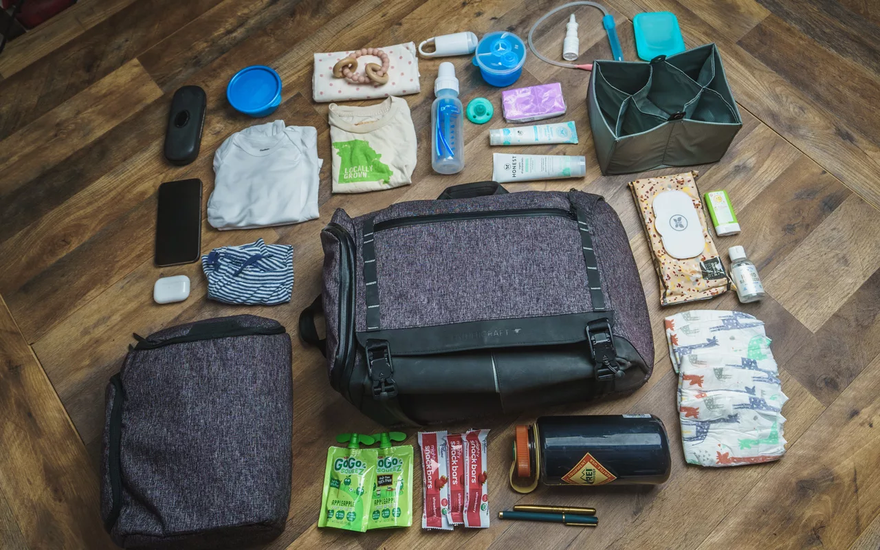 The contents of a diaper bag laid out on a table with a diaper bag for dads