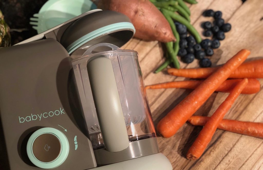 The Babycook on a counter with carrots, a sweet potato, and blueberries